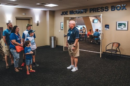 A group of people in blue shirts stands in front of the Joe McGuff Press Box, listening to a guide.