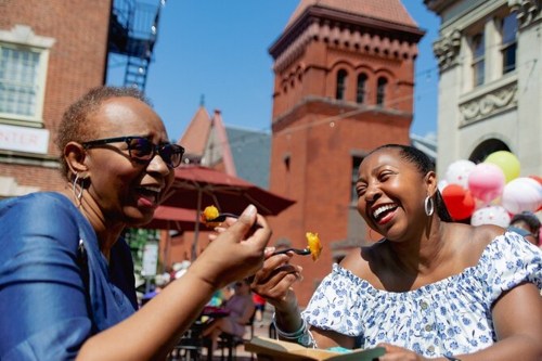 Two women laughing and enjoying food outdoors, with colorful balloons and historic buildings in the background.