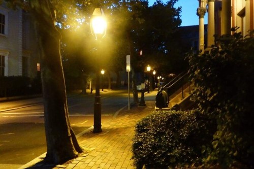 A quiet street at dusk, lined with trees and vintage street lamps casting a warm glow on the brick sidewalk.