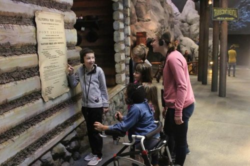 A group of children gathers around a sign in a rustic setting, listening intently to a guide.