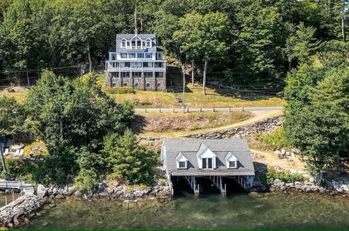 Aerial view of a large house on a hillside next to a waterfront boathouse surrounded by trees.