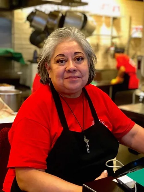 A woman in a red shirt and black apron smiles at the camera, working in a kitchen setting.