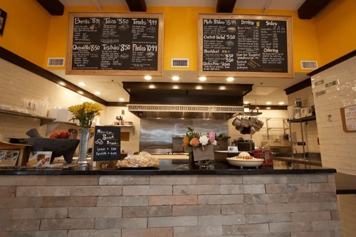 A cozy café interior featuring a menu board, counter with flowers, and various food items displayed.