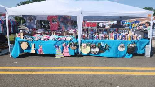 A colorful market stall with clothing and crafts displayed under a white tent on a sunny day.