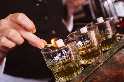 A hand lights a sugar cube on fire above glasses of green liquid, creating a dramatic effect in a bar setting.