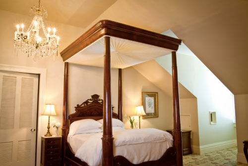 A cozy bedroom featuring a four-poster bed, chandelier, and soft lighting, with sloped ceiling and elegant decor.
