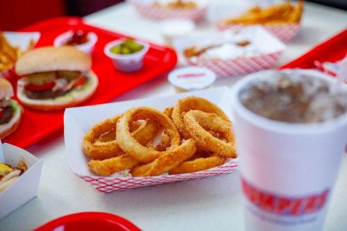 A close-up of crispy onion rings on a tray, with burgers, fries, and a drink in the background.
