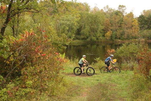 Two cyclists ride along a grassy path near a calm lake, surrounded by trees and autumn foliage.