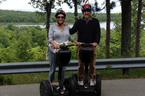 A couple stands on Segways with a scenic view of trees and a river in the background, both wearing helmets and smiling.