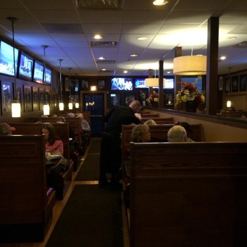 A dimly lit restaurant interior with wooden booths, diners, and a waiter attending to customers. TVs are visible on the walls.
