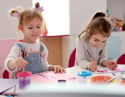 Two young girls are painting at a table, focused on their artwork with colorful supplies around them.