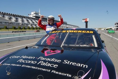 A race car driver celebrates with raised arms on top of a stock car at a racetrack.