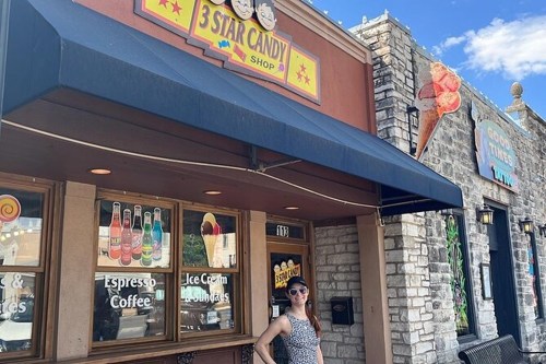 A woman stands outside the 3 Star Candy Shop, featuring a colorful sign and a stone building backdrop.