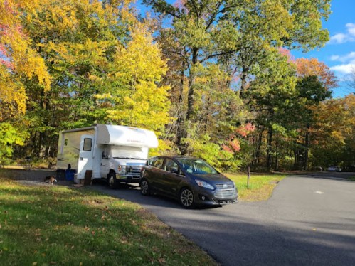 A parked RV and car in a scenic area surrounded by colorful autumn trees.