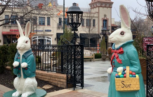 Two large, colorful rabbit statues in a park setting, one holding a basket and the other wearing a coat.