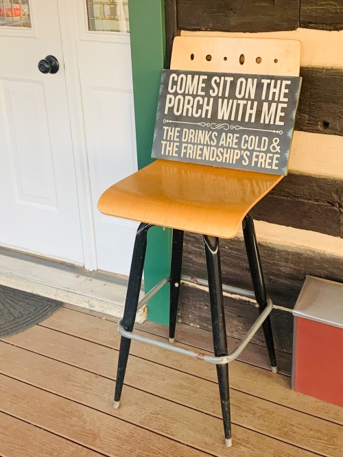 A wooden stool on a porch holding a sign that invites people to sit and enjoy cold drinks and friendship.