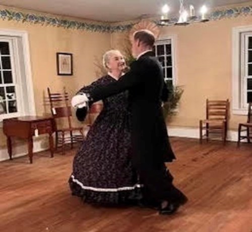 A man and woman in vintage attire dance together in a warmly lit room with wooden floors and simple decor.