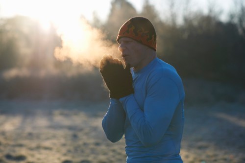 A man in a blue long-sleeve shirt and a beanie exhales visible breath in a frosty outdoor setting.
