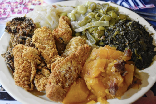 A plate of Southern-style food featuring fried fish, greens, beans, cabbage, and sweet potatoes.