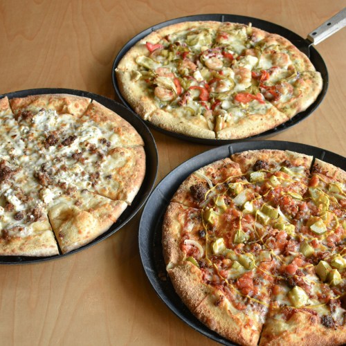 Three pizzas on black trays: one with meat and cheese, one with vegetables, and one with a mix of toppings.