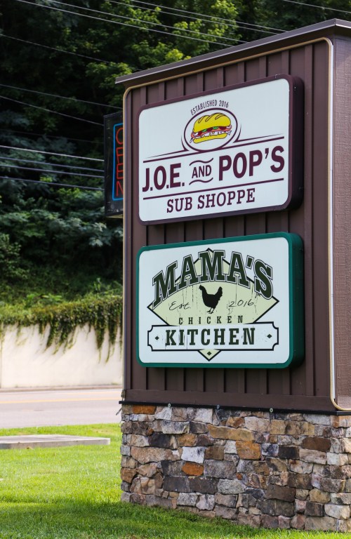 Sign for J.O.E. and Pop's Sub Shoppe and Mama's Chicken Kitchen, featuring colorful logos and a stone base.