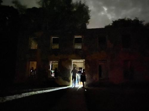 A group of people stands outside a dark, abandoned building, illuminated by flashlights under a cloudy night sky.