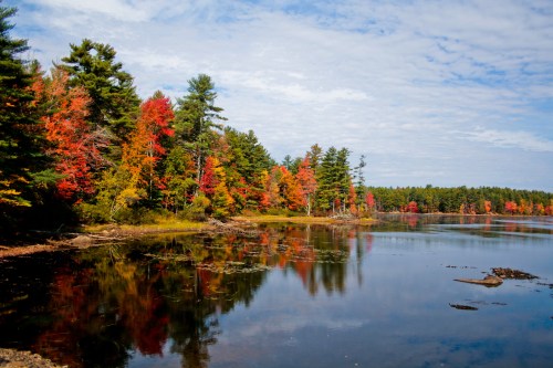A serene lake surrounded by vibrant autumn trees reflecting in the water under a partly cloudy sky.