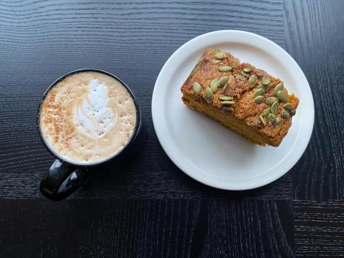 A black coffee cup with latte art beside a slice of cake topped with pumpkin seeds on a white plate.