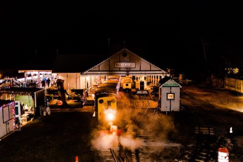 A brightly lit train station at night, with a train emitting steam and colorful lights decorating the buildings.