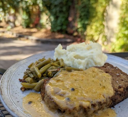 A plate of meatloaf with gravy, mashed potatoes, and green beans, set outdoors with greenery in the background.