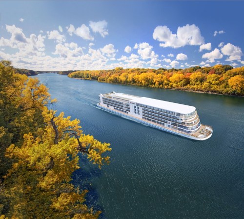 A modern river cruise ship glides through a scenic waterway surrounded by vibrant autumn foliage and a clear blue sky.