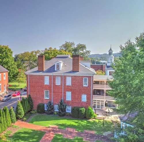 A historic brick building surrounded by greenery, with trees and a distant church steeple visible in the background.