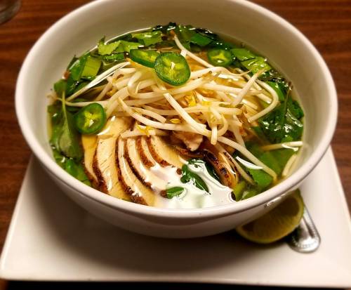 A bowl of clear broth soup with sliced chicken, bean sprouts, herbs, and lime on the side.