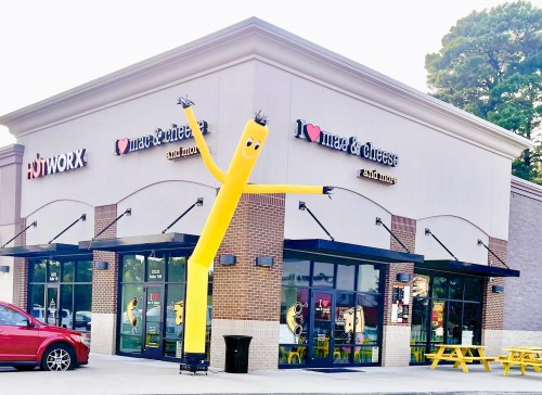 A restaurant with a yellow inflatable dancer outside, featuring signs for "I Love Mac & Cheese" and "Hotworx."