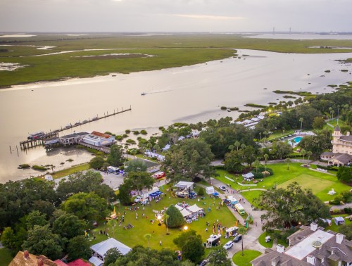 Aerial view of a waterfront festival with tents, greenery, and a calm river in the background under a cloudy sky.