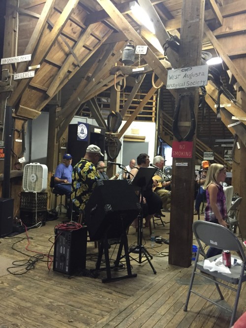 A band performs in a rustic wooden hall with signs, fans, and audience members enjoying the music.