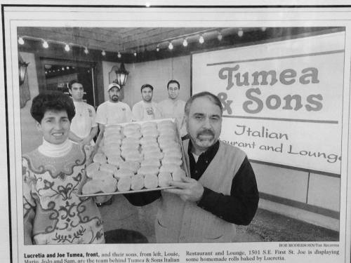 A black and white photo of a family outside Tumea & Sons restaurant, holding a tray of homemade rolls.