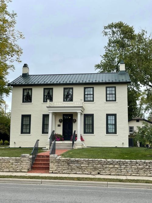 A two-story white house with a black metal roof, surrounded by greenery and a stone wall, on a cloudy day.