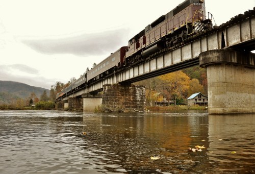 A train crosses a bridge over a river, surrounded by autumn foliage and mountains under a cloudy sky.