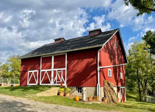 A red barn with white trim sits on a grassy area, surrounded by trees and pumpkins under a partly cloudy sky.