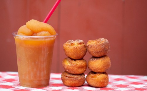A cup of slushy drink next to a stack of five mini cinnamon sugar donuts on a checkered tablecloth.