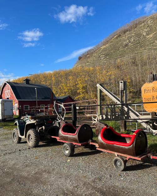 A red barn and a mountain backdrop with a cart for a ride, parked next to an ATV on a gravel path.