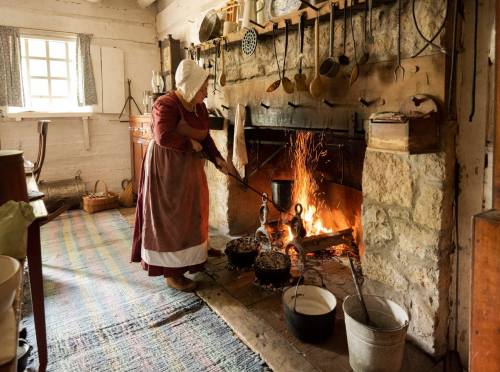 A person in historical attire stirs a pot over a roaring fire in a rustic kitchen with stone walls and wooden utensils.