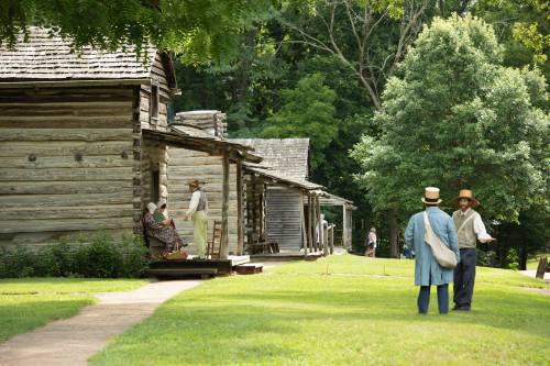 Two men in period clothing converse outside rustic log cabins, with trees and people in the background.