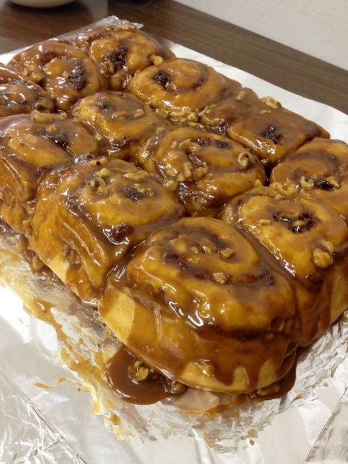 A tray of freshly baked cinnamon rolls topped with caramel and nuts, glistening on a silver foil surface.