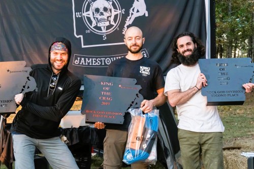 Three men proudly hold awards at an outdoor event, standing in front of a black banner with a skull logo.