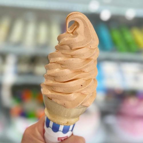 A hand holding a swirl of soft serve ice cream in a cone, with colorful ice cream options blurred in the background.