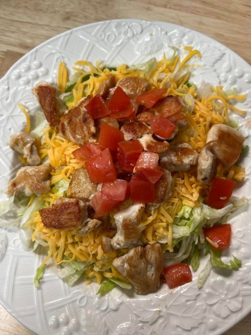 A plate of salad topped with shredded cheese, diced tomatoes, and pieces of chicken on a bed of lettuce.