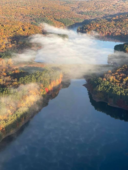 Aerial view of a river surrounded by colorful autumn foliage and mist rising from the water.
