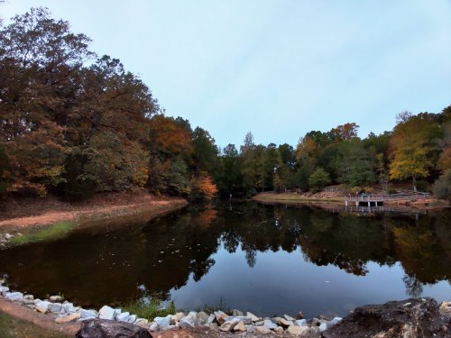 A serene pond surrounded by trees with autumn foliage, reflecting the sky and landscape in calm water.